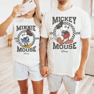 Puede incluir: Dos camisetas blanquecinas con gr&aacute;ficos a juego con Mickey y Minnie Mouse. Las camisetas tienen un dise&ntilde;o vintage con los personajes dentro de un c&iacute;rculo a cuadros, el texto "Mickey Mouse" y "Minnie Mouse", y el logotipo de Disney.