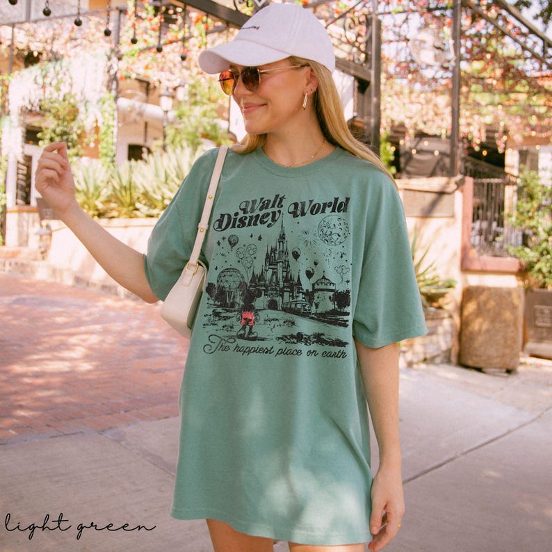 Puede incluir: Camiseta verde claro con un gr&aacute;fico del castillo de Walt Disney World, Mickey Mouse y el texto "The happiest place on earth". La camiseta se lleva con una gorra blanca y gafas de sol.
