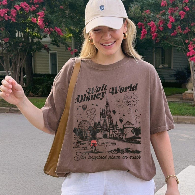 Puede incluir: Camiseta marr&oacute;n con un gr&aacute;fico del castillo de Cenicienta, fuegos artificiales y el texto "Walt Disney World" y "The happiest place on earth". La camiseta se usa con una gorra de b&eacute;isbol beige y pantalones cortos blancos.