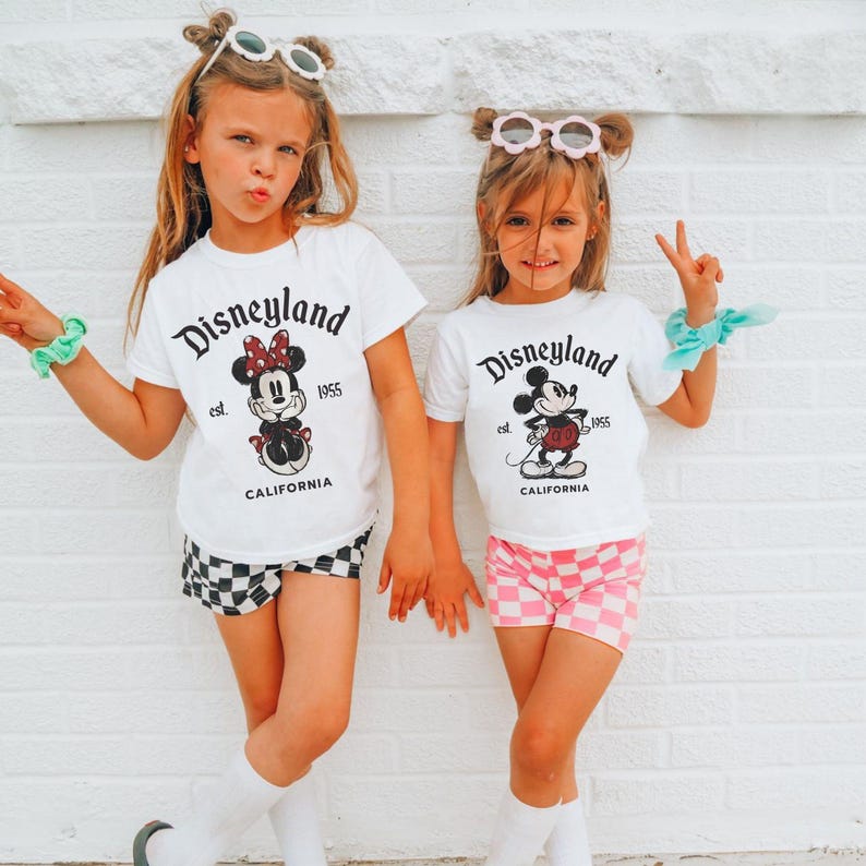 May include: Two young girls wearing matching white t-shirts with a Disneyland graphic, black and white checkered shorts, and pink checkered shorts. The shirts feature Mickey and Minnie Mouse designs and the text "Disneyland est. 1955 California."