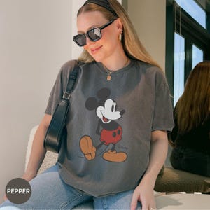 Puede incluir: Una camiseta gris con un dise&ntilde;o de Mickey Mouse. El personaje de dibujos animados se muestra en su pose cl&aacute;sica, con pantalones cortos rojos y zapatos marrones. La camiseta tiene un aspecto vintage y descolorido.