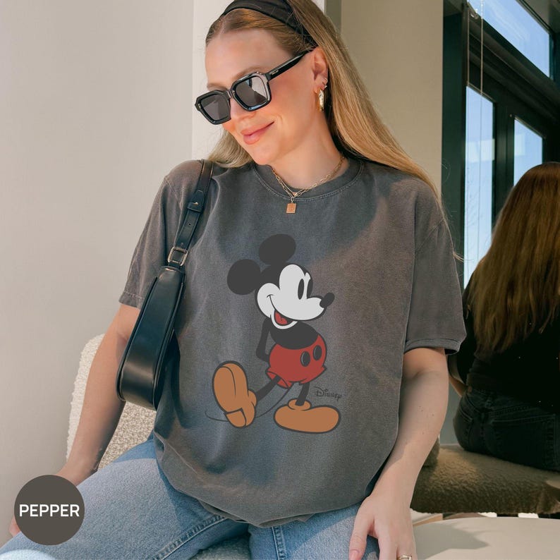 Puede incluir: Una camiseta gris con un dise&ntilde;o de Mickey Mouse. El personaje de dibujos animados se muestra en su pose cl&aacute;sica, con pantalones cortos rojos y zapatos marrones. La camiseta tiene un aspecto vintage y descolorido.