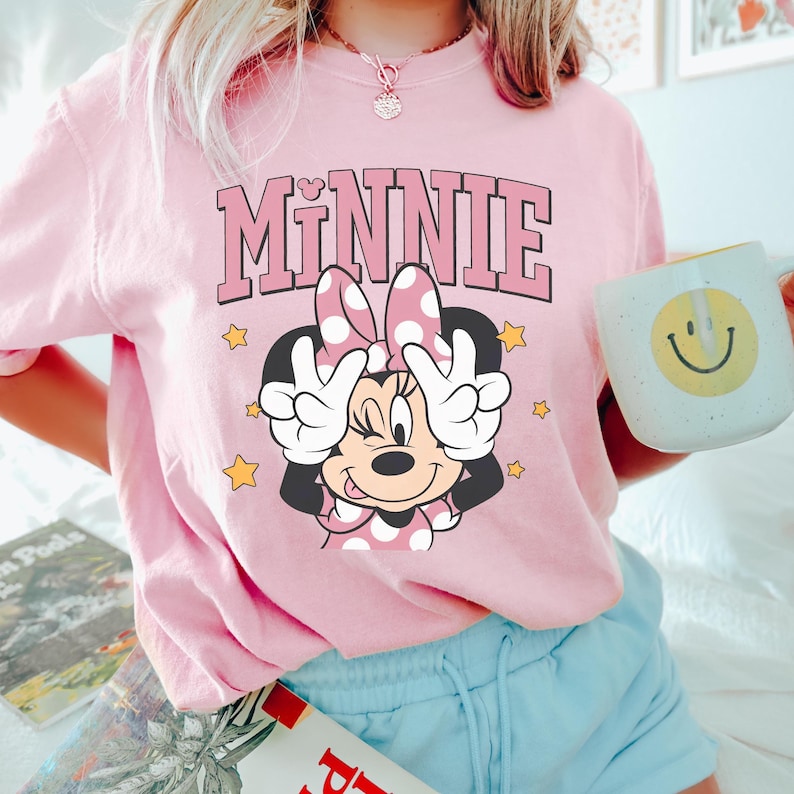 以下が含まれることがあります： ミニーマウスのグラフィックが特徴の薄いピンク色のTシャツ。「MINNIE」の文字がピンク色のブロック体でプリントされ、ミニーマウスがピースサインをしているイラストの上に配置されています。黄色の星がキャラクターを囲んでいます。黄色のスマイリーフェイスの白いマグカップが手に持たれています。