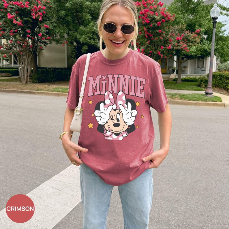 以下が含まれることがあります： ピンク色の文字で「MINNIE」と書かれたクリムゾン色のTシャツ。水玉模様のリボンとピースサインをしたミニーマウスのグラフィック。明るいブルーのジーンズと合わせています。