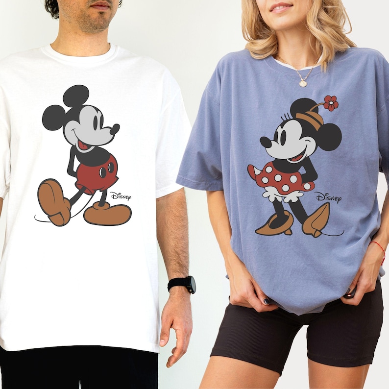 Puede incluir: Dos camisetas con personajes de Disney. Una es blanca con Mickey Mouse con pantalones cortos rojos y zapatos marrones. La otra es azul con Minnie Mouse con falda de lunares rojos y zapatos marrones. Ambas tienen el logo de Disney.