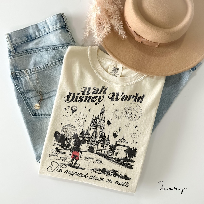 May include: Ivory-colored t-shirt with a black graphic of Mickey Mouse in front of a castle, with the words "Walt Disney World" and "The happiest place on earth." The shirt is paired with light blue jeans, a tan hat, and a necklace.
