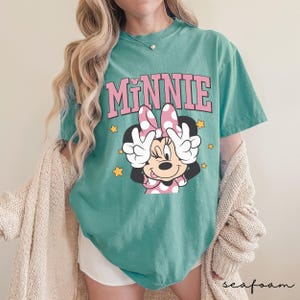 以下が含まれることがあります： ピンク色の文字で「MINNIE」と書かれた、ミニーマウスのグラフィックが特徴のティール色のTシャツ。ミニーはピンクのドット柄のリボンをつけ、ピースサインをしています。黄色の星がグラフィックの周りに散りばめられています。