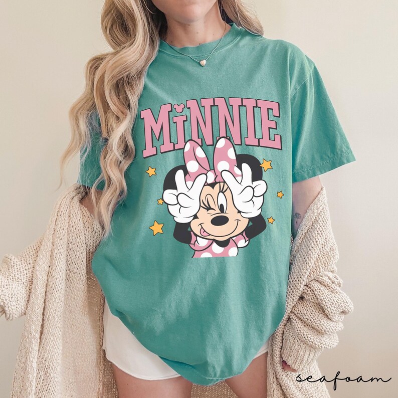 以下が含まれることがあります： ピンク色の文字で「MINNIE」と書かれた、ミニーマウスのグラフィックが特徴のティール色のTシャツ。ミニーはピンクのドット柄のリボンをつけ、ピースサインをしています。黄色の星がグラフィックの周りに散りばめられています。