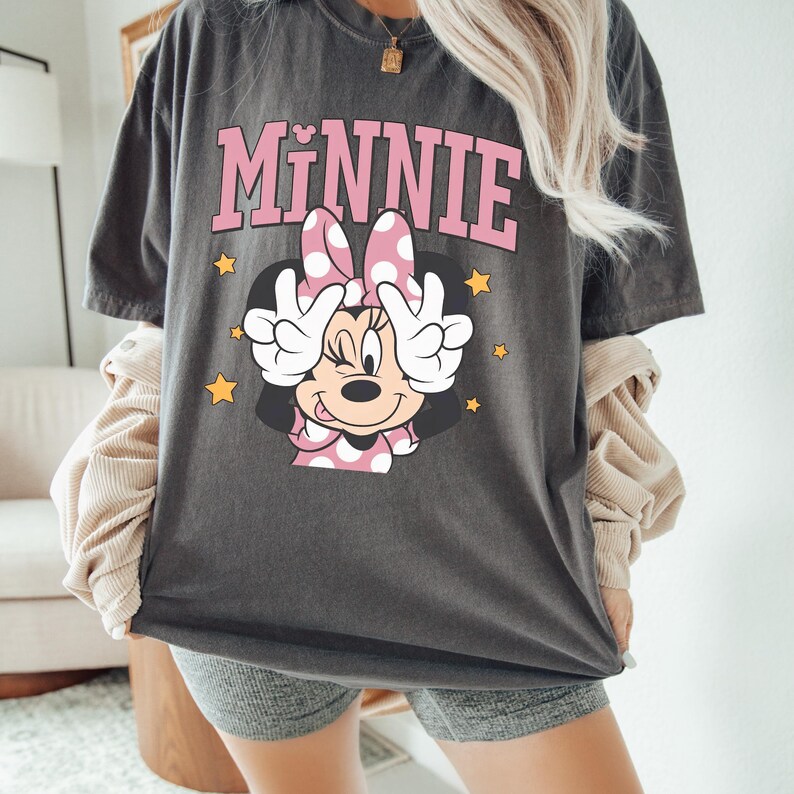 以下が含まれることがあります： ピンク色の「MINNIE」の文字がプリントされた濃いグレーのTシャツ。ミニーマウスの漫画のキャラクターが描かれており、ピンクのドット柄のリボンとドレスを着ています。ミニーマウスは両手でピースサインをしており、周りには黄色の星が描かれています。