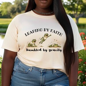 May include: A cream-colored t-shirt featuring a graphic of three green frogs in white shirts. The text reads LEAPING BY FAITH above the frogs and humbled by gravity below them. The frogs are shown jumping and landing on the ground with small stars.