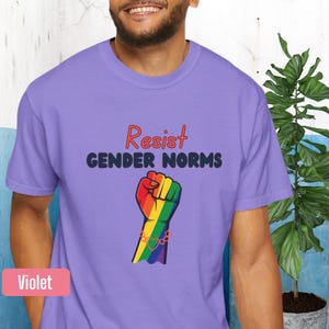 Può includere: Una t-shirt viola con la scritta rossa Resist e la scritta nera GENDER NORMS sopra un'immagine di un pugno alzato con strisce arcobaleno e il sottile contorno di un braccialetto arancione a forma di cuore sul polso.