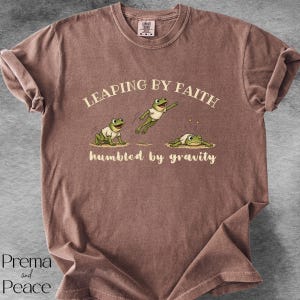 May include: A brown short-sleeve t-shirt featuring a graphic of three green frogs in white shirts. The text reads LEAPING BY FAITH above the frogs and humbled by gravity below them. The shirt has a crew neckline and a visible brand label at the neck.