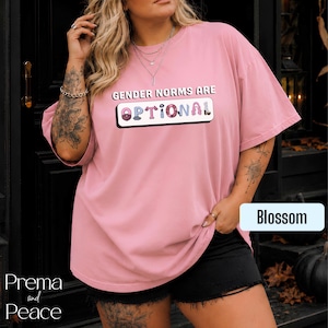 May include: A dusty pink short-sleeve t-shirt featuring the text GENDER NORMS ARE OPTIONAL in black and white lettering. The word OPTIONAL is stylized with various colorful patterns inside each letter. The shirt has a crew neckline and loose fit.
