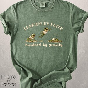 May include: A sage green short-sleeve t-shirt featuring a graphic of three frogs in white t-shirts. The text reads LEAPING BY FAITH above the frogs and humbled by gravity below them. The design shows one frog sitting, one jumping, and one landing.