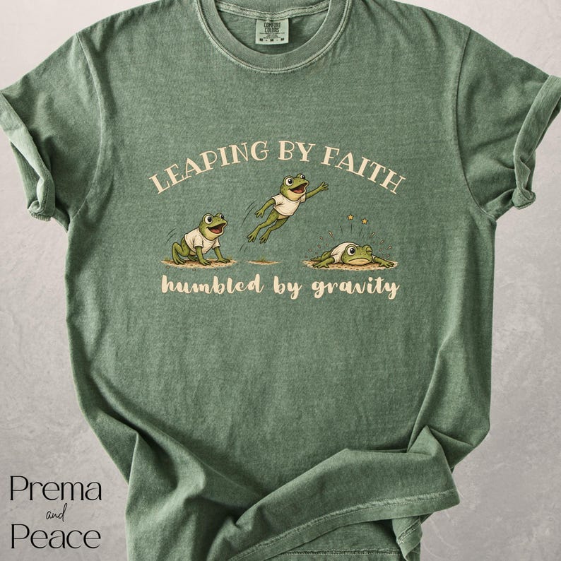 May include: A sage green short-sleeve t-shirt featuring a graphic of three frogs in white t-shirts. The text reads LEAPING BY FAITH above the frogs and humbled by gravity below them. The design shows one frog sitting, one jumping, and one landing.
