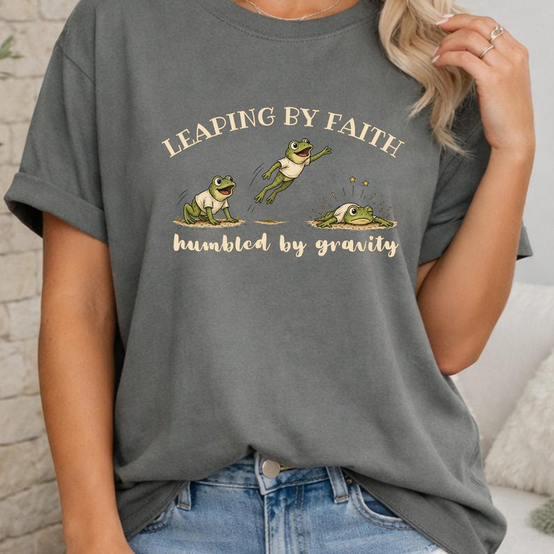May include: A charcoal gray t-shirt featuring a graphic of three green frogs in white shirts. The text reads LEAPING BY FAITH above the frogs and humbled by gravity below them. The middle frog is mid-air, while the third frog is on the ground.