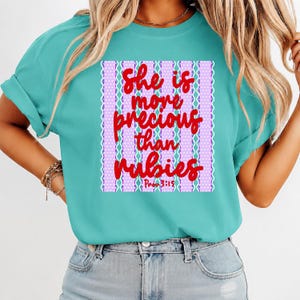 Op de afbeelding: Turquoise T-shirt met de rode tekst "She is more precious than rubies". De tekst staat tegen een paars-witte achtergrond met patroon. Het shirt heeft korte mouwen en een ronde hals.