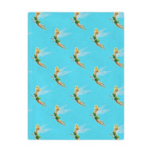 May include: A light blue blanket with a repeating pattern of a cartoon fairy with blonde hair, a green dress, and translucent wings. The fairy is in a flying pose. The blanket has a soft texture.