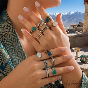 Op de afbeelding: Een collectie ringen met diverse ontwerpen en edelstenen, waaronder smaragdgroene, blauwe en witte stenen. De ringen worden op een hand getoond, waarbij verschillende stijlen en metaalafwerkingen worden uitgelicht. De achtergrond toont een schilderachtig uitzicht op bergen en een gebouw.
