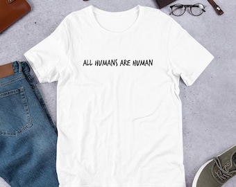 May include: A white t-shirt with the black text "ALL HUMANS ARE HUMAN". The shirt is on a light grey surface, with blue jeans, a brown wallet, a watch, and shoes visible.