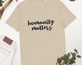 May include: A beige t-shirt featuring the words "humanity matters" in black cursive lettering. The shirt has a crew neck and is made of a soft fabric. The image also includes a pair of jeans, a wooden branch, and a beaded bracelet.