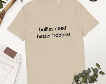 May include: A beige t-shirt with the slogan "bullies need better hobbies" printed in black. The tee shirt is made of a soft material and is likely a unisex design. The shirt is displayed on a neutral background.