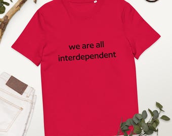 May include: A red t-shirt with the text "we are all interdependent" in black. The shirt has a crew neck and is made of a soft material. The image also shows a pair of white jeans, a wooden branch, and a beaded necklace.