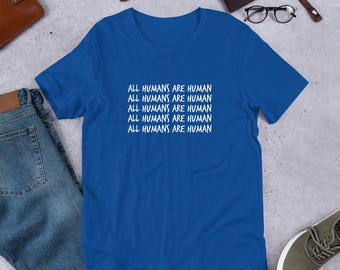 May include: Royal blue t-shirt with the white text "ALL HUMANS ARE HUMAN" repeated five times. The shirt is displayed with blue jeans, a brown wallet, a watch, and a pair of glasses. The shirt is a casual wear item.