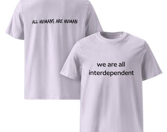 May include: Two light purple t-shirts. One shirt has the text "ALL HUMANS ARE HUMAN" on the back. The other shirt has the text "we are all interdependent" on the front. The shirts are made of a soft material.
