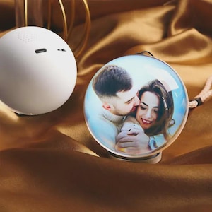 Peut inclure: Un haut-parleur Bluetooth sphérique blanc et un ornement photo rond et transparent. L'ornement affiche un couple s'embrassant. Le haut-parleur a un port de charge et des trous de haut-parleur. Le fond est un tissu doré-brun brillant.