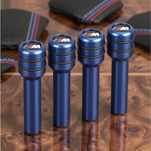 May include: Four blue cylindrical door lock pins featuring the M logo on the top cap. Each pin has two black rubber O-rings around the upper section. The metallic blue finish is smooth, and the M logo consists of a blue, violet, and red stripe.