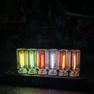 May include: A row of six small, clear glass cylindrical vials, each containing a vertical glowing filament. The filaments emit distinct colors including yellow, green, red, white, orange, and a faint light, all mounted on a black base.