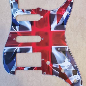 May include: A guitar pickguard featuring a stylized Union Jack flag design in red, white, and blue. The plastic plate includes three cutouts for pickups, a slot for a switch, and several small pre-drilled holes for mounting screws around the perimeter.