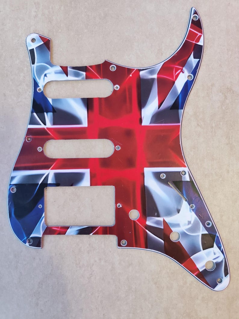 May include: A guitar pickguard featuring a stylized Union Jack flag design in red, white, and blue. The plastic plate includes three cutouts for pickups, a slot for a switch, and several small pre-drilled holes for mounting screws around the perimeter.