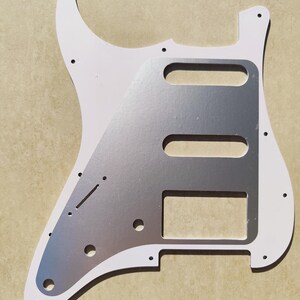 May include: A white electric guitar pickguard featuring a metallic silver center plate. The pickguard has pre-cut openings for three pickups, a switch slot, and holes for volume and tone control knobs, along with several small mounting screw holes.