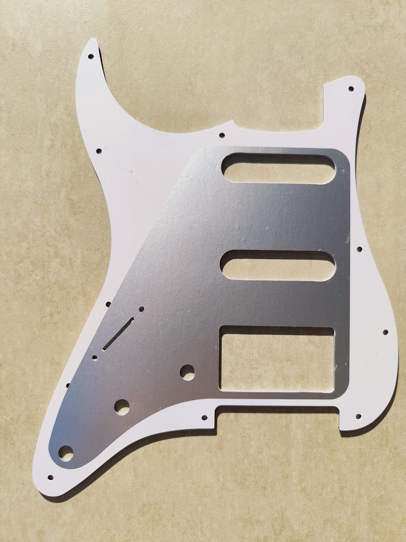 May include: A white electric guitar pickguard featuring a metallic silver center plate. The pickguard has pre-cut openings for three pickups, a switch slot, and holes for volume and tone control knobs, along with several small mounting screw holes.