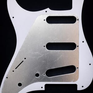 Puede incluir: Golpeador de guitarra blanco con una placa de metal plateado. El golpeador tiene una forma curva con varios orificios para tornillos y recortes para las pastillas y los controles. La placa de metal tiene tres largas aberturas rectangulares.