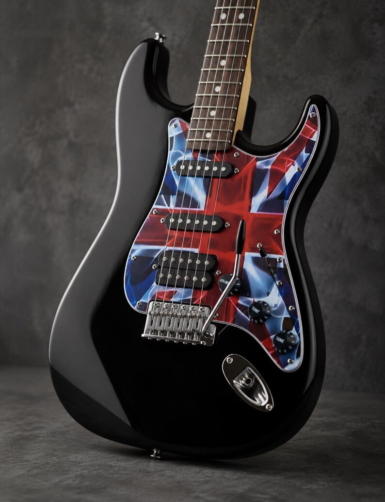 May include: A black electric guitar featuring a pickguard with a red, white, and blue Union Jack flag pattern. The instrument includes three black pickups, a chrome bridge, a tremolo arm, and two black control knobs on the glossy black body.