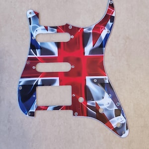 May include: A guitar pickguard featuring a stylized Union Jack flag pattern in red, white, and blue. The plastic plate includes cutouts for three pickups, a volume knob, two tone knobs, and a five-way selector switch, along with multiple screw holes.