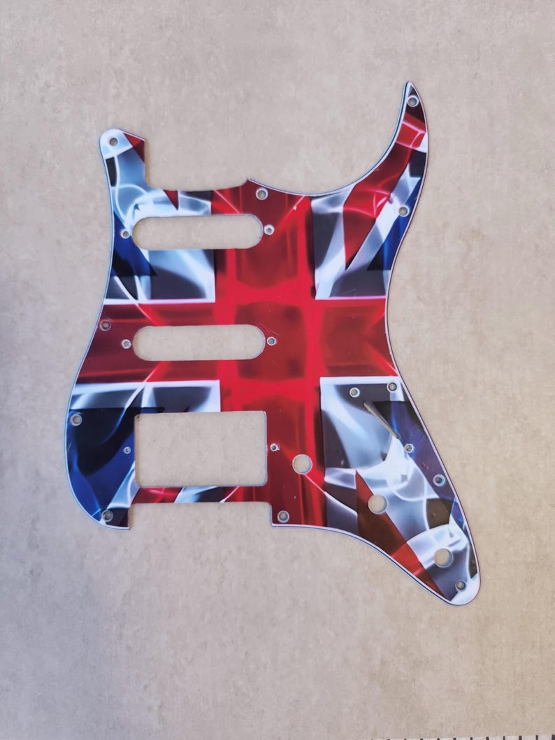 May include: A guitar pickguard featuring a stylized Union Jack flag pattern in red, white, and blue. The plastic plate includes cutouts for three pickups, a volume knob, two tone knobs, and a five-way selector switch, along with multiple screw holes.