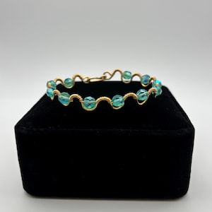 Peut inclure: Un bracelet en fil doré orné de perles bleu-vert iridescentes. Les perles sont espacées uniformément et fixées avec un design en fil délicat et tissé. Le bracelet est présenté sur une surface en velours noir.