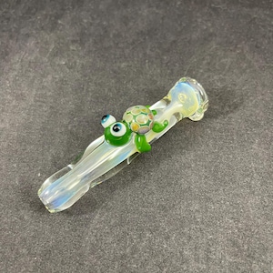 May include: A clear glass pipe with a turtle design. The turtle is green with a patterned shell and large blue eyes. The pipe has a flared bowl and a long, slender stem with iridescent coloring.