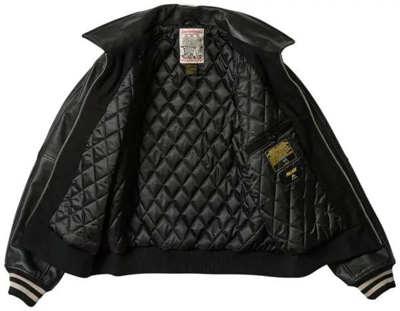 May include: Black leather jacket with a quilted interior. The jacket has a wide collar, a ribbed hem, and cuffs with white and tan stripes. The interior features a pocket with a gold label. The jacket is open, displaying the interior.