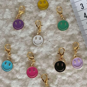 May include: A selection of smiley face charms in a variety of colours, each with a gold-coloured clasp. The charms feature a simple smiley face design and are available in yellow, purple, green, white, blue, pink, black, and lavender.