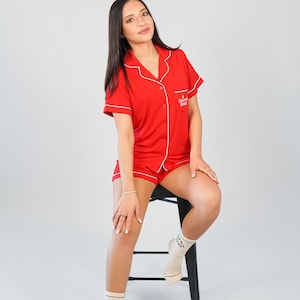 Pu&ograve; includere: Completo pigiama rosso con rifiniture bianche, composto da una camicia a maniche corte con bottoni e pantaloncini abbinati. La camicia ha il testo "Dormi Bene" ricamato sul petto. La persona indossa calzini con un disegno verde.