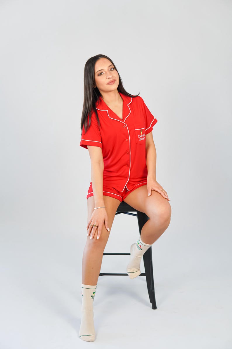 Pu&ograve; includere: Completo pigiama rosso con rifiniture bianche, composto da una camicia a maniche corte con bottoni e pantaloncini abbinati. La camicia ha il testo "Dormi Bene" ricamato sul petto. La persona indossa calzini con un disegno verde.