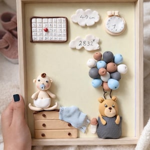 Puede incluir: Una obra de arte enmarcada con temática de bebé con un fondo beige claro. Presenta un calendario, un reloj, una figura de bebé, un oso de peluche y un grupo de globos. También se incluye el texto "49 cm" y "2832".