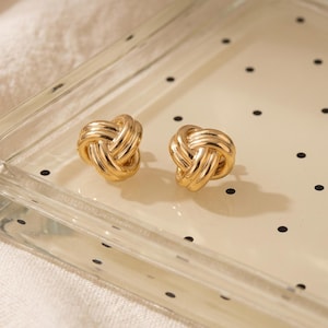 May include: A pair of gold-toned stud earrings featuring a polished, intertwined knot design. Each earring consists of multiple smooth, rounded metal strands woven together into a compact, circular shape with a reflective, metallic finish.