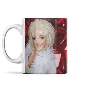 May include: White ceramic mug featuring a smiling person with blonde hair, wearing a white feathered boa and a pearl necklace. The background is a red, gem-like pattern. The mug has a white handle.