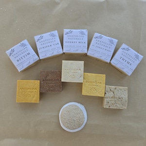 May include: A collection of five Anatolian Naturals handmade soap bars in paper packaging labeled Bittim, Juniper Tar, Donkey Milk, St. John's Wort, and Thyme, alongside five unwrapped square soap bars and a round, beige, textured loofah sponge.